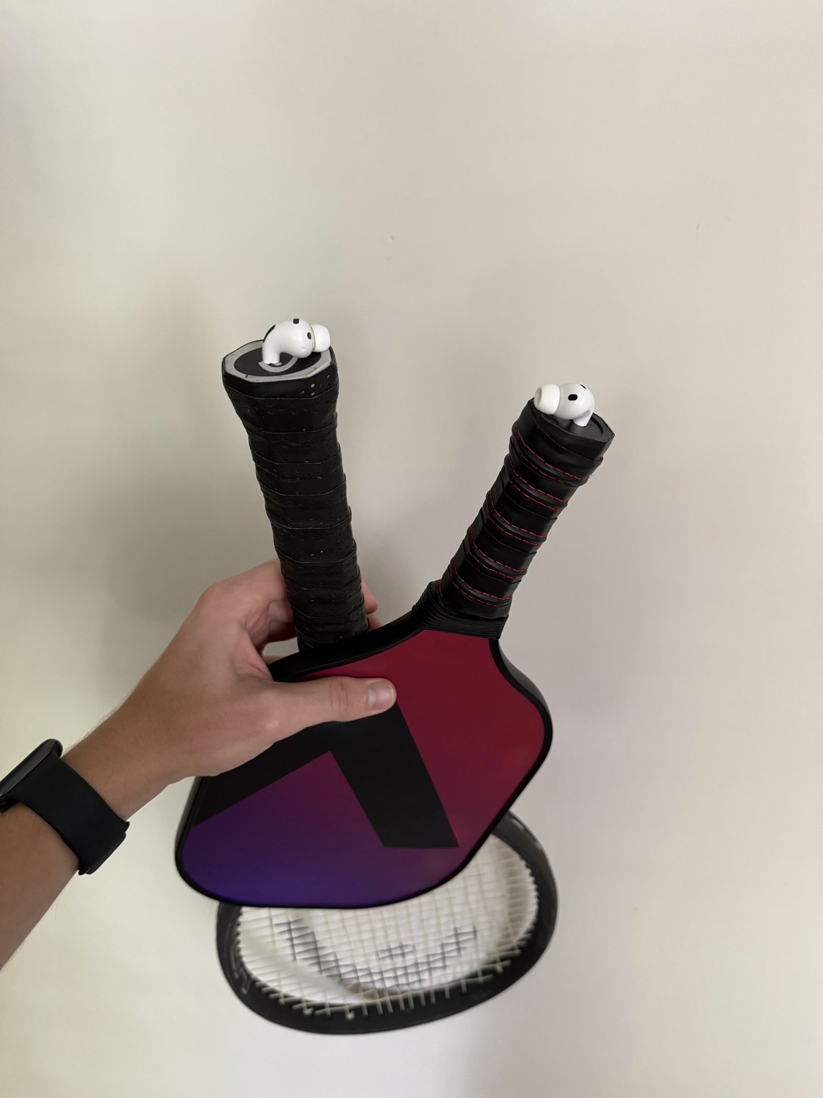 A pickleball paddle and a tennis racket, each with an AirPod tucked into a hole in the end of the grip